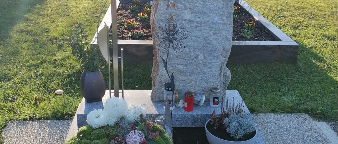 Grab mit Steinrohling als Grabstein, Metallkreuz, Blumen und Dekoration.