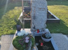 Grab mit Steinrohling als Grabstein, Metallkreuz, Blumen und Dekoration.