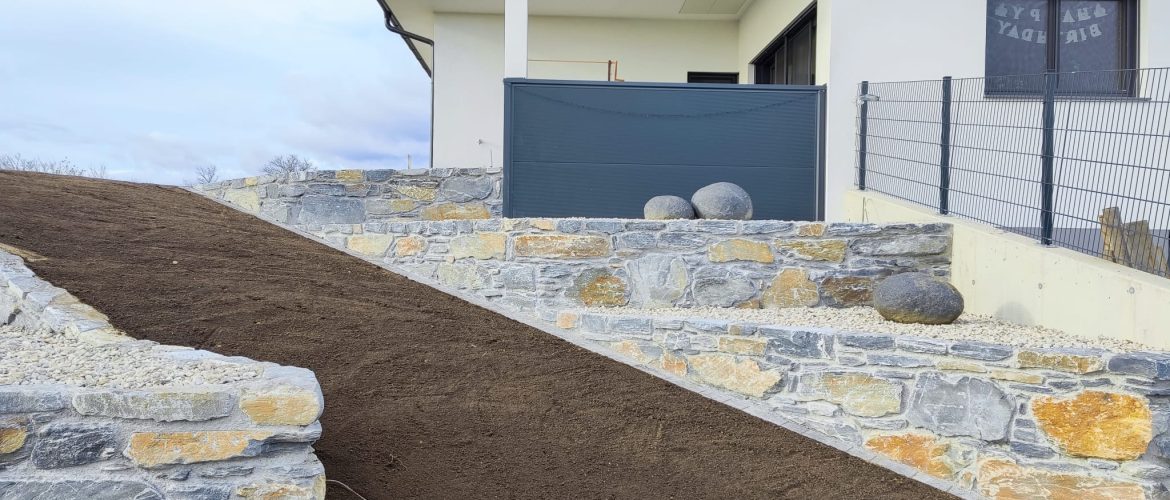 Schiefersteinmauer mit integrierter Rampe, neben einem weißen, modernen Wohnhaus.