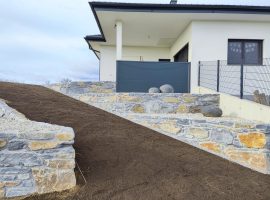 Schiefersteinmauer mit integrierter Rampe, neben einem weißen, modernen Wohnhaus.