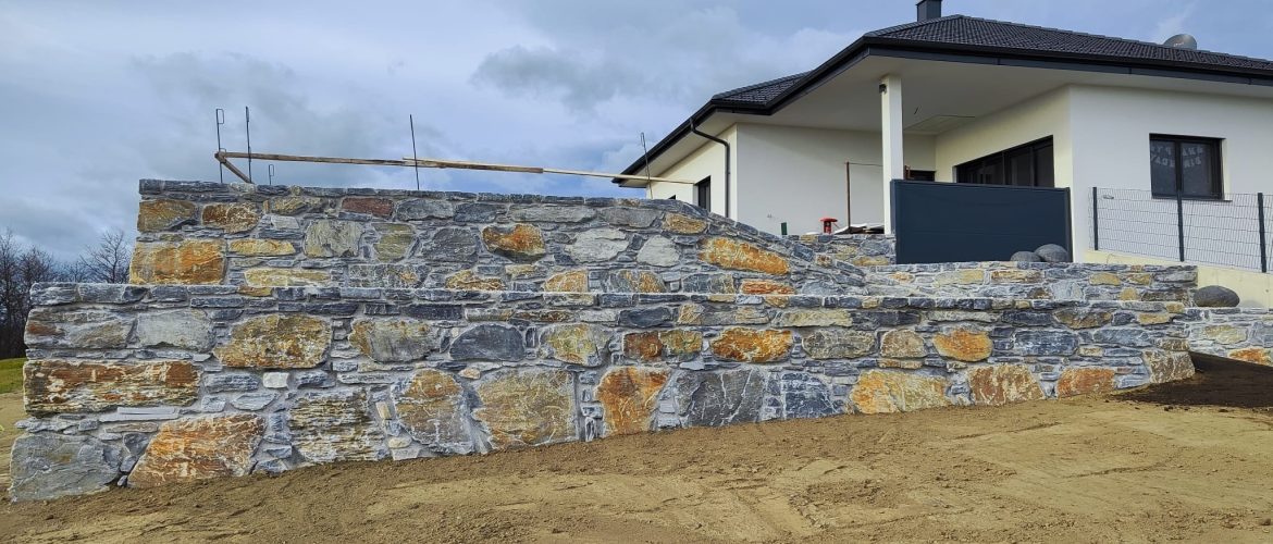 Etagenförmige Schiefermauer mit verschiedenen Ebenen und moderner Architektur im Hintergrund.