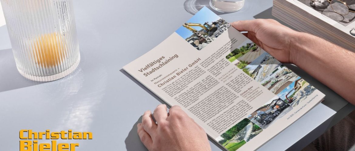 Person liest einen Artikel über die Christian Bieler GmbH im Gemeinde-Kurier Stadtschlaining, mit Bildern von Natursteinmauern und Erdarbeiten. Auf dem Tisch stehen ein Glas Wasser, eine Zeitschrift und eine Lampe mit Zitrone im Glas.